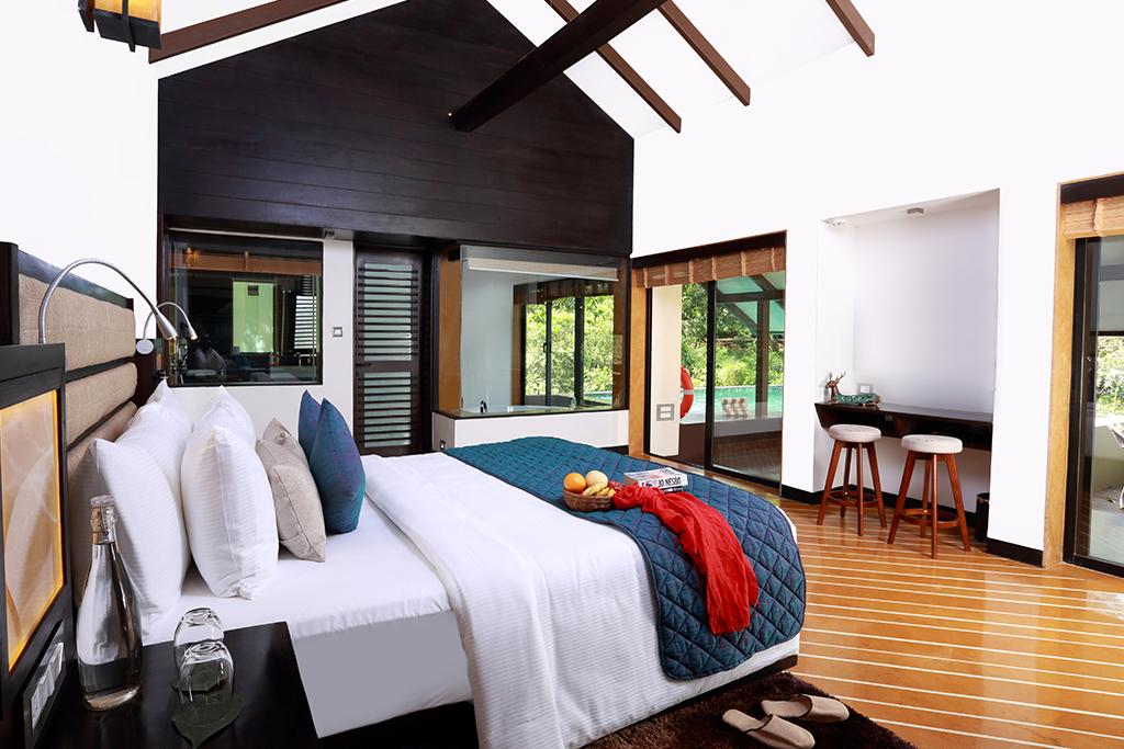 Dressed up bed with white sheets and pillows and blue cushions and bed covering in a room with bay windows showcasing a private pool and the forest outside.